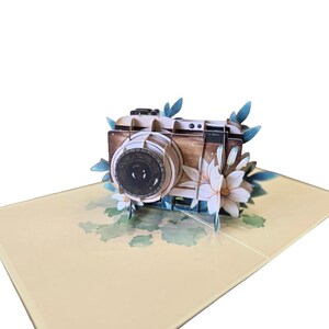 Peut inclure: Carte de vœux pop-up 3D avec un appareil photo vintage. L'appareil photo est marron et beige, orné de fleurs blanches et de feuilles bleues. La carte repose sur une surface jaune clair avec des motifs floraux à l'aquarelle.