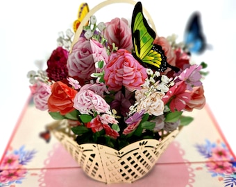 May include: A 3D pop-up card featuring a basket of pink, red, and white flowers with green leaves. A green butterfly with black markings is perched on the flowers. The card is open and the flowers are in focus.