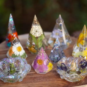 May include: A collection of clear resin ring holders in various shapes, including cones and flower-like bowls, each embedded with colorful dried flowers, green foliage, and shimmering glitter accents throughout the transparent material.