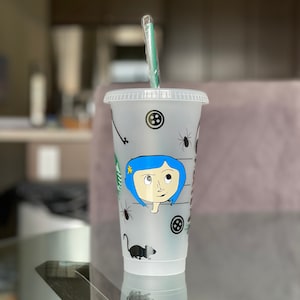 Pode incluir: Um copo frio de pl&aacute;stico transparente com tampa e canudo verde. O copo apresenta uma ilustra&ccedil;&atilde;o de desenho animado de uma menina com cabelo azul, bot&otilde;es, aranhas e um rato. O logotipo da Starbucks &eacute; vis&iacute;vel.
