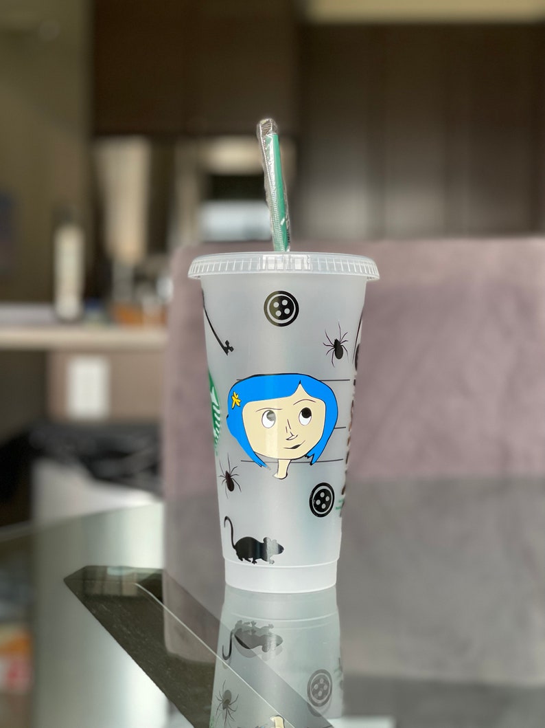 Pode incluir: Um copo frio de pl&aacute;stico transparente com tampa e canudo verde. O copo apresenta uma ilustra&ccedil;&atilde;o de desenho animado de uma menina com cabelo azul, bot&otilde;es, aranhas e um rato. O logotipo da Starbucks &eacute; vis&iacute;vel.