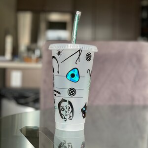 Pode incluir: Um copo de pl&aacute;stico transparente para bebidas frias com tampa e canudo verde. O copo &eacute; decorado com gr&aacute;ficos pretos e azuis de uma aranha, bot&otilde;es e rostos de personagens. O copo est&aacute; sobre uma superf&iacute;cie de vidro.