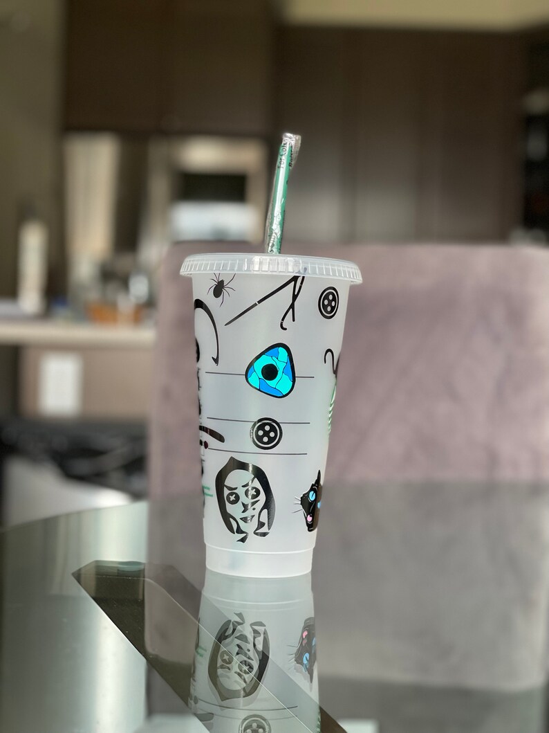 Pode incluir: Um copo de pl&aacute;stico transparente para bebidas frias com tampa e canudo verde. O copo &eacute; decorado com gr&aacute;ficos pretos e azuis de uma aranha, bot&otilde;es e rostos de personagens. O copo est&aacute; sobre uma superf&iacute;cie de vidro.