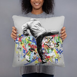 May include: A gray throw pillow with a black and white illustration of a person kicking a wall covered in colorful graffiti. The graffiti includes various shapes, lines, and letters.