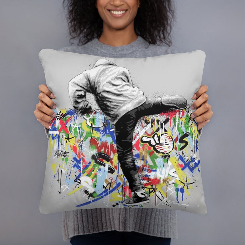 May include: A gray throw pillow with a black and white illustration of a person kicking a wall covered in colorful graffiti. The graffiti includes various shapes, lines, and letters.
