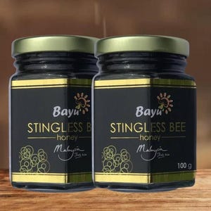 以下が含まれることがあります： Bayu Stingless Beeの蜂蜜が入ったガラス瓶が2つ。黒いラベルには金色の装飾とブランド名が施されています。瓶には「蜂蜜」と「マレーシア」と書かれています。各瓶には100gの蜂蜜が入っています。