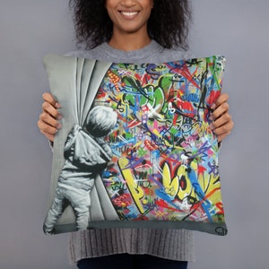 May include: A colorful throw pillow featuring a child peeking around a corner, revealing a wall covered in vibrant graffiti. The graffiti includes a variety of colors, shapes, and letters, creating a dynamic and energetic design.