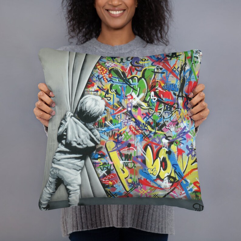 May include: A colorful throw pillow featuring a child peeking around a corner, revealing a wall covered in vibrant graffiti. The graffiti includes a variety of colors, shapes, and letters, creating a dynamic and energetic design.