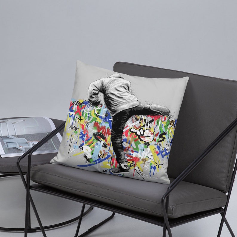 May include: A throw pillow with a white background featuring a black and white illustration of a person kicking a wall covered in colorful graffiti.
