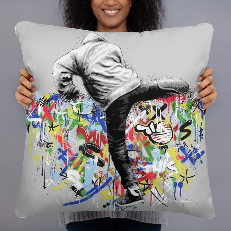 May include: A gray throw pillow with a colorful graffiti design featuring a figure in a white hoodie kicking a wall. The figure is wearing black pants and sneakers. The graffiti is in various colors, including red, blue, green, yellow, and orange.