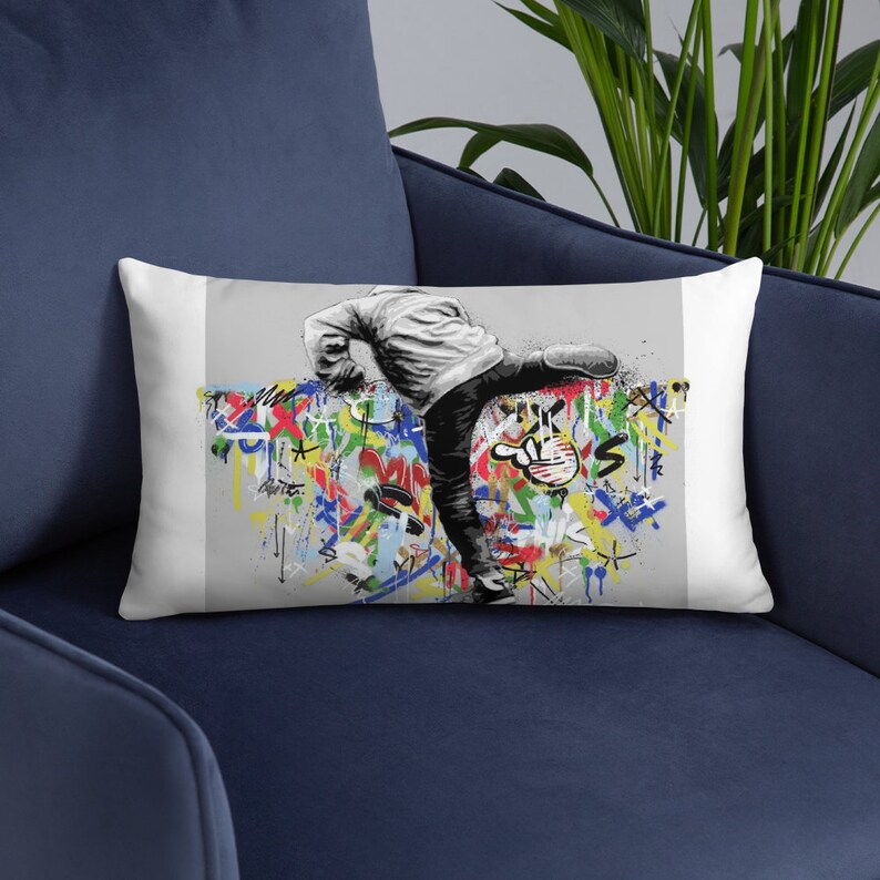 May include: A white throw pillow with a colorful graffiti design featuring a person kicking a wall. The person is wearing a white jacket and jeans.