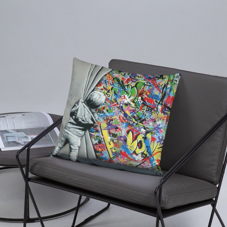 May include: A throw pillow featuring a colorful graffiti background with a child peeking out from behind a white curtain. The child is wearing a grey hooded sweatshirt and pants. The graffiti includes the word "LOVE" in large yellow letters.