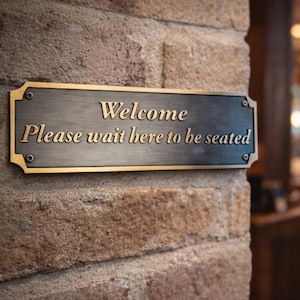 Pode incluir: Uma placa retangular de metal com as palavras "Welcome Please wait here to be seated" em letras douradas. A placa está montada em uma parede de tijolos texturizados, marrom claro. O fundo está desfocado, mostrando um interior de restaurante.