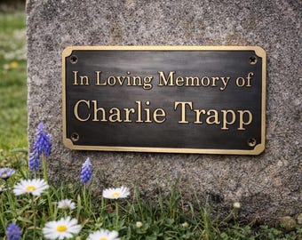 May include: A rectangular bronze memorial plaque with the inscription "In Loving Memory of Charlie Trapp." The plaque is mounted on a weathered stone surface, surrounded by grass, daisies, and bluebell flowers.