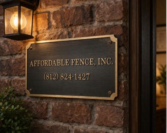 May include: A rectangular dark metal plaque with gold-colored lettering reading AFFORDABLE FENCE, INC. and the phone number (812) 824-1427, mounted on a rustic red brick wall next to a black outdoor wall-mounted lantern light fixture.