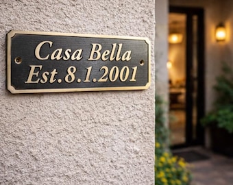 May include: A rectangular black plaque with a gold-colored border and gold-colored raised lettering that reads Casa Bella Est. 8.1.2001. The plaque features two visible screw holes on the left and right sides for mounting purposes.