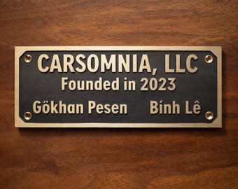 May include: A rectangular brass plaque with the text "CARSOMNIA, LLC" and "Founded in 2023" in gold lettering. The names "Gökhan Pesen" and "Bình Lê" are also inscribed. The plaque is mounted on a dark wooden surface.