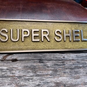 May include: A rectangular brass sign with the words "SUPER SHELL" in raised, silver-colored letters. The sign has a textured, golden-brown background and is mounted on a weathered wooden surface. Two screw holes are visible.