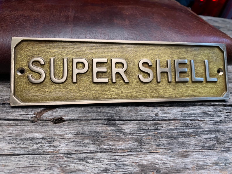 May include: A rectangular brass sign with the words "SUPER SHELL" in raised, silver-colored letters. The sign has a textured, golden-brown background and is mounted on a weathered wooden surface. Two screw holes are visible.