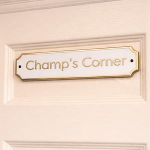 Może przedstawiać: Prostokątny biały znak ze złotą obwódką i napisem "Champ's Corner" złotymi literami. Znak jest zamontowany na białych drzwiach z zagłębionym wzorem panelu. Czarne kropki znajdują się na każdym końcu znaku.