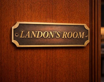 Puede incluir: Un letrero de puerta rectangular de metal con acabado marrón oscuro y borde dorado. El letrero presenta el texto LANDON'S ROOM en letras mayúsculas doradas y está fijado a una puerta de madera con dos tornillos visibles en los lados.