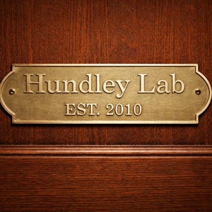 May include: A brass sign with a curved top and bottom is fixed to a dark wooden door. The sign reads "Hundley Lab" in a serif font, with "EST. 2010" below. The sign is attached with two small screws.