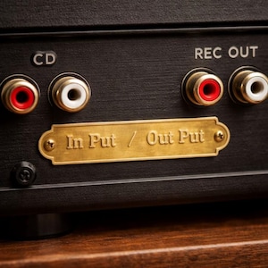 May include: Close-up of the back panel of a black audio device. It features gold-colored input/output ports labeled "CD" and "REC OUT." A gold plate reads "In Put / Out Put."