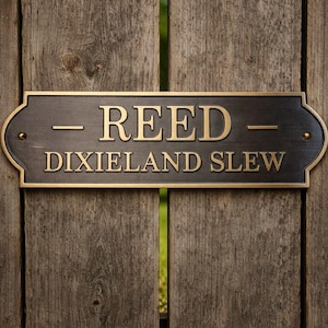 Puede incluir: Un letrero rectangular de metal marrón oscuro con letras y borde dorados está montado en una valla de madera desgastada. El letrero dice "REED DIXIELAND SLEW" en una fuente serif.