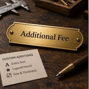 May include: A brass plaque with the words "Additional Fee" engraved in a serif font. A small card with the words "Custom Additions" and icons for extra text, logo/artwork, and size/thickness is next to the plaque. A wooden pen is also visible.