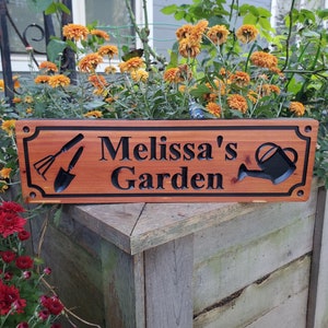 May include: A wooden sign with the text "Melissa's Garden" carved into it. The sign is painted brown and has black lettering. There are two gardening tools and a watering can carved into the sign.