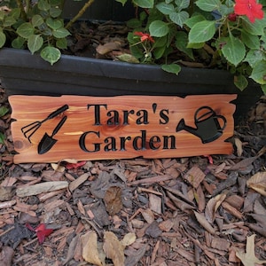 May include: A wooden sign with the text "Tara's Garden" carved into it. The sign also features a carved image of a watering can and gardening tools.