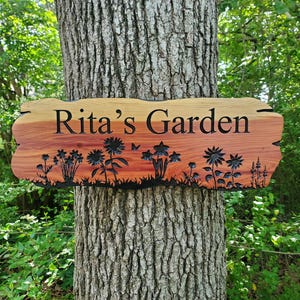 May include: A rectangular wooden sign with a natural live edge, featuring the engraved text "Rita's Garden" in black lettering. The wood displays a reddish-brown grain, and the bottom section is decorated with black silhouettes of various flowers.