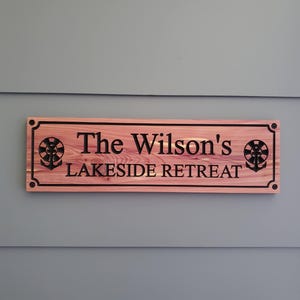 May include: A rectangular wooden sign with a reddish-brown wood grain finish featuring black engraved text that reads The Wilson's LAKESIDE RETREAT. The sign includes black anchor icons on both sides and is mounted with four black corner screws.