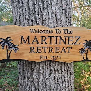 May include: Wooden sign with the text "Welcome To The MARTINEZ RETREAT Est. 2025" and palm tree silhouettes. The sign is attached to a tree trunk. The sign has a natural wood grain finish with black lettering.