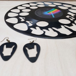 May include: A pair of black guitar pick earrings with a rock and roll hand gesture cut out. The earrings are hanging from a light brown surface next to a black vinyl record with a similar design.
