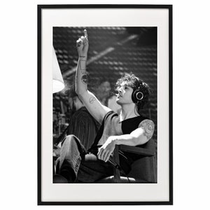 May include: Black and white framed print featuring a person with curly hair wearing headphones and a two-tone shirt, seated in a chair. The person is raising their arm, pointing upwards. Tattoos are visible on the arm. The print is in a black frame.