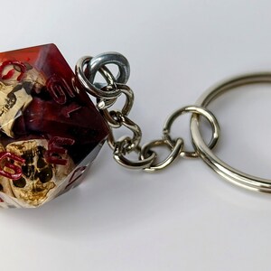 May include: A red and clear resin dice keychain with a skull design. The dice is attached to a silver metal keychain.