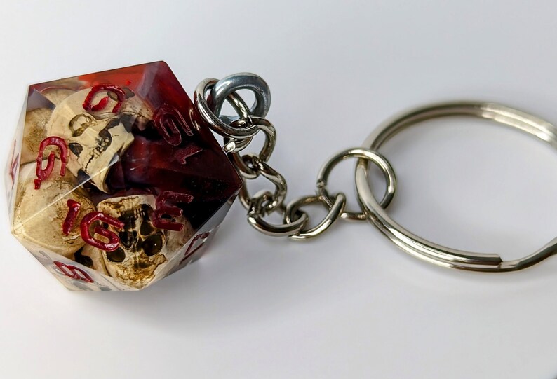 May include: A red and clear resin dice keychain with a skull design. The dice is attached to a silver metal keychain.