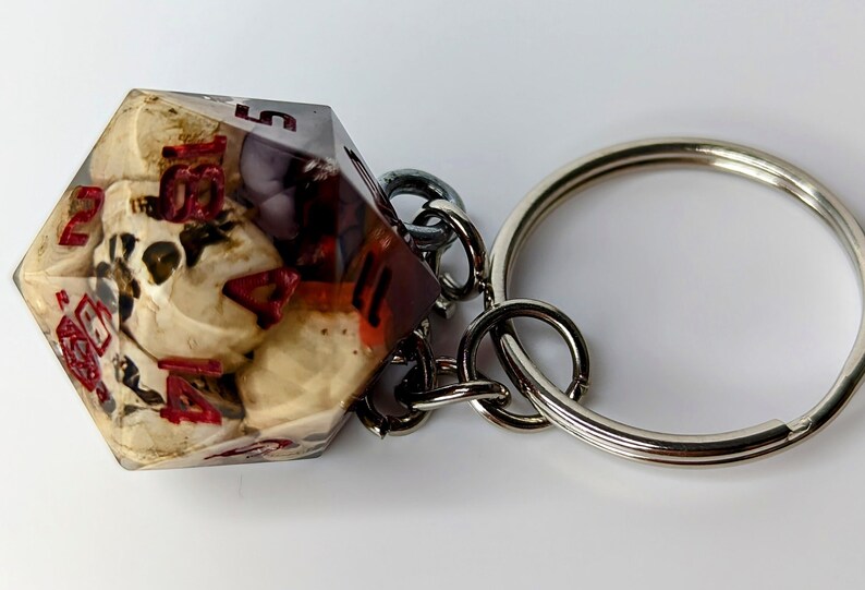 May include: A white and red resin dice keychain with a silver key ring. The dice has a skull design inside.