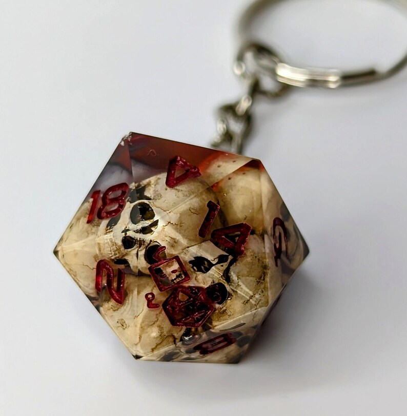 May include: A clear resin dice with a skull design and red numbers. The dice is attached to a silver keychain.