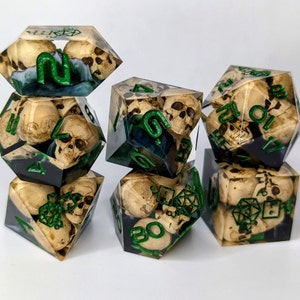 May include: Six green and white dice with skull and bone designs. The dice are made of clear resin and have a green and white swirl pattern. The dice are numbered 1-6.