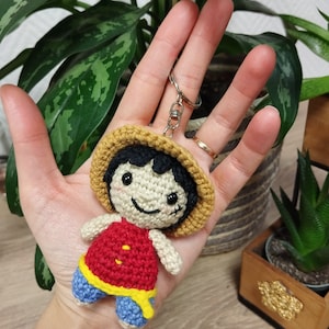 Peut inclure: Un porte-clés crocheté fait main représentant un personnage de dessin animé avec un chapeau de paille, un haut rouge, un short bleu et des cheveux noirs. Le porte-clés est attaché à un anneau en métal argenté. Le personnage a un visage souriant.