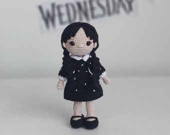 May include: A crocheted doll with black hair, a black dress with white polka dots, and black shoes. The doll is standing on a white surface.
