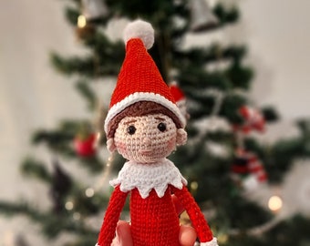 May include: A crocheted elf doll wearing a red and white hat and a red and white outfit. The elf is being held in a person's hand in front of a Christmas tree.