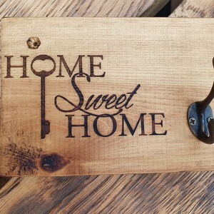 May include: Wooden sign with the text "Home Sweet Home" and a keyhole design. A metal hook is attached to the right side of the sign.