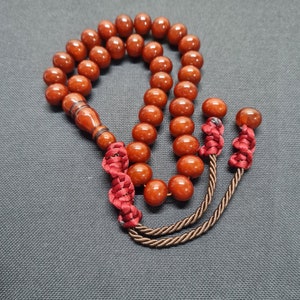 Könnte beinhalten: Eine Gebetskette mit großen, runden, rotbraunen Perlen. Die Perlen sind mit einer dunkelroten Kordel und einem dekorativen Holzelement aufgefädelt. Die Quaste besteht aus einer gedrehten braunen Kordel und roten Knoten.