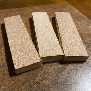 May include: Three rectangular sanding blocks with a brown, textured surface. The blocks are made of wood and are approximately 6 inches long.