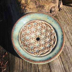 Könnte beinhalten: Eine runde Keramikschale mit einem erhabenen, geprägten geometrischen Lebensblumenmuster in der Mitte. Die Schale hat ein hellcremefarbenes Inneres mit einem blau-grün glasierten Rand und einem kleinen kreisförmigen Loch in der Mitte.