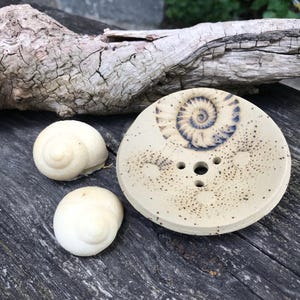 Könnte beinhalten: Eine beige Keramik-Seifenschale mit blau-weißem Spiralmuster, zusammen mit zwei weißen, schneckenförmigen Seifen. Die Schale hat drei Abflusslöcher und eine gesprenkelte Oberfläche. Auf einer verwitterten Holzoberfläche angeordnet.