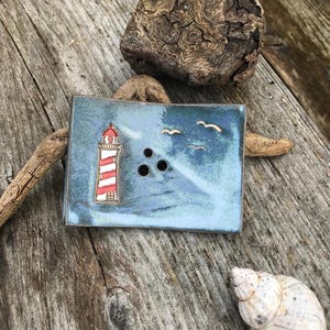 Könnte beinhalten: Ein rechteckiger Knopf mit glänzender blauer Emaille-Oberfläche, der auf der linken Seite eine Illustration eines rot-weiß gestreiften Leuchtturms und in der Mitte drei kleine kreisförmige Löcher sowie drei kleine metallische Vogelsilhouetten zeigt.